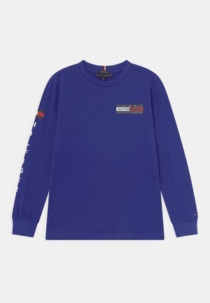 Blå langærmet bluse med rund halsudskæring med "Tommy Hilfiger Outdoors"-logo på brystet, hvid tekst på højre ærme og et lille flag på venstre manchet.
