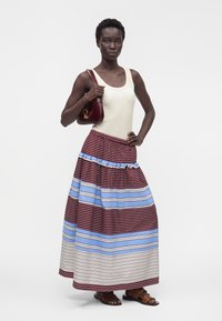 Femme portant un haut blanc sans manches, une longue jupe rayée bordeaux, bleu et blanc, des sandales marron, tenant un petit sac à main rouge foncé.
