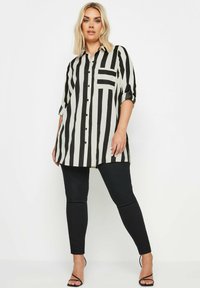 Camicia a righe bianco e nero con design a bottoni, maniche lunghe, tasca frontale e orlo curvo, abbinata a pantaloni neri aderenti.