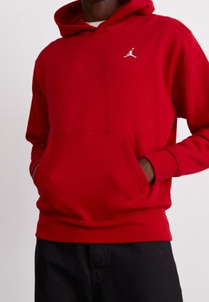 Osoba nosząca czerwoną bluzę z kapturem, z rękami w przedniej kieszeni i małym białym logo Jumpman na klatce piersiowej, w połączeniu z czarnymi spodniami.
