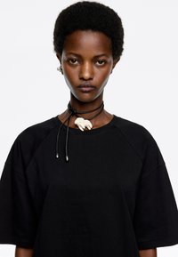 T-shirt nera oversize con maniche corte, impreziosita da un choker in pelle nera decorato con un piccolo pendente a forma di elefante bianco.
