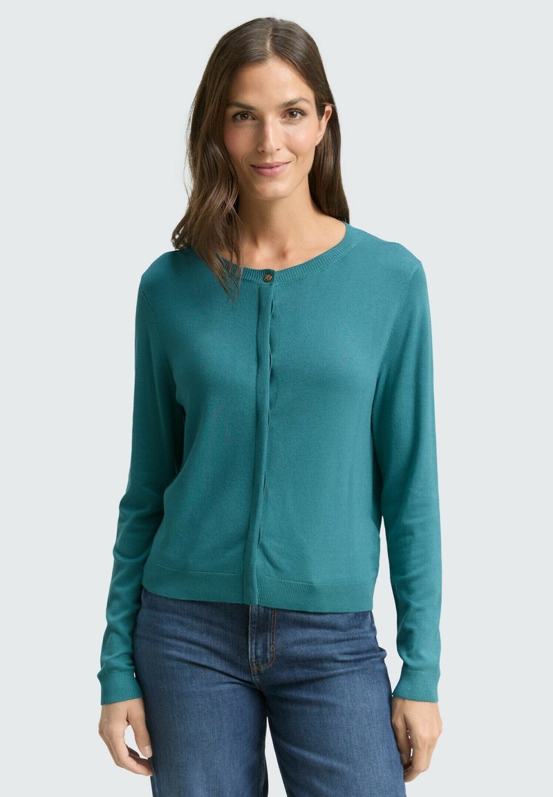 Turkusowy kardigan z długimi rękawami, okrągłym dekoltem i pojedynczym drewnianym guzikiem, wykonany z miękkiej dzianiny. Stylizowany z dżinsami.