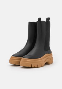 Zwarte enkelboots met een leren bovenwerk, elastische zijpanelen en dikke tan rubberen zolen met een golvend profiel en loopvlakpatroon.