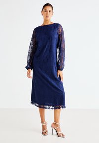 Vestido de renda azul-marinho com mangas longas e transparentes, apresentando padrões florais intrincados. Comprimento midi com uma silhueta reta. Combinado com saltos finos.