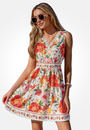 Kobieta w białej sukience bez rękawów z czerwonym i pomarańczowym nadrukiem kwiatów i ptaków, trzymająca rąbek sukienki, w okularach przeciwsłonecznych i z długimi falującymi włosami.