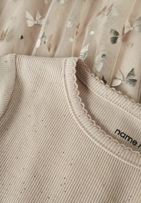 Haut beige clair côtelé avec un décolleté festonné, doté d'une texture douce et de paillettes métalliques, associé à une superposition transparente à motifs de papillons.