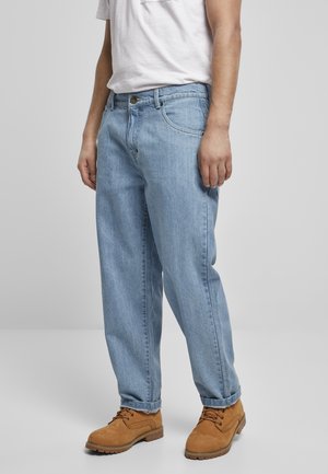 Homme portant un jean en denim bleu clair, des bottes lacées couleur fauve et un t-shirt blanc, debout devant un fond gris uni.