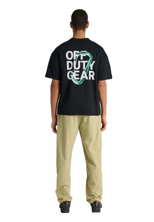 Mann trägt ein schwarzes T-Shirt mit dem Schriftzug "OFF DUTY GEAR" und einem grünen Karabiner-Grafik auf dem Rücken, beige Hose und schwarze Schuhe, steht mit dem Rücken zum Betrachter.