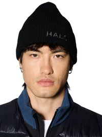 Jonge man die een zwarte gebreide muts draagt met de tekst "HALO.", zilveren oorringen en een donker jack met een blauwe kraag, dichtgeritst.