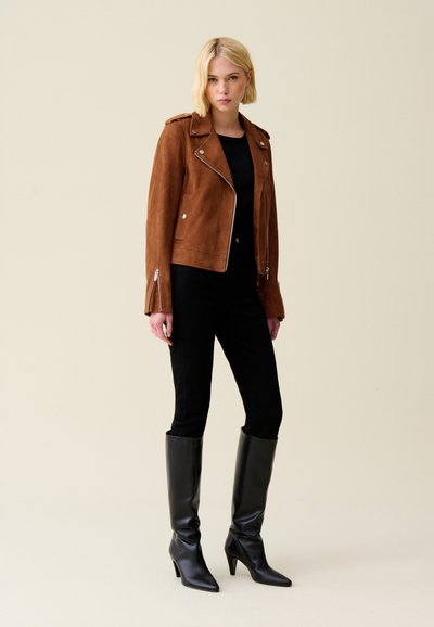 Giacca da biker in suede marrone con tasche con zip e hardware argentato, abbinata a pantaloni neri attillati e stivali neri con tacco alto.