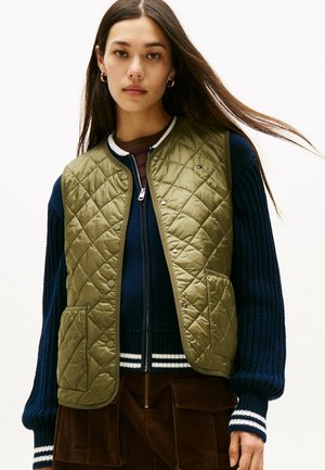 Gilet trapuntato verde oliva con chiusure a pressione e tasche laterali, indossato sopra un maglione a coste blu navy con polsini a righe e una chiusura con zip.