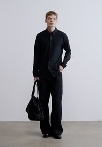 Fekete hosszú ujjú ing gombokkal, sötét, széles szárú nadrággal párosítva. A személy egy fekete bevásárlótáskát tart, semleges színű padlón állva.