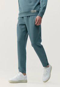 Tyrkysové tepláky vyrobené z mäkkého materiálu, zúžený strih, bočné vrecká a štítok na páse. Kombinované s bielymi teniskami.