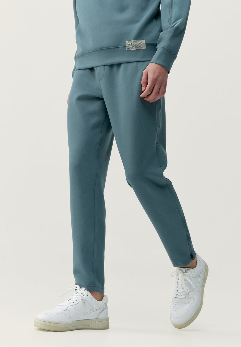 Tyrkysové tepláky vyrobené z mäkkého materiálu, zúžený strih, bočné vrecká a štítok na páse. Kombinované s bielymi teniskami.