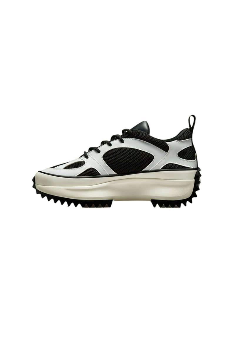 Schwarzer und weißer Sneaker aus Netz- und glattem Leder, mit einer dicken Sohle, die mit spitzen Noppen versehen ist, und einem Zugriemen an der Ferse.