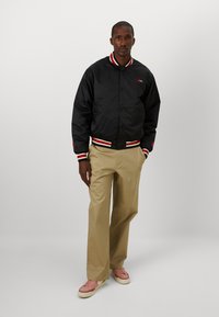 Black bomber jacket with red and white striped ribbed cuffs and hem. Paired with khaki pants and light pink sneakers, standing against a plain background.