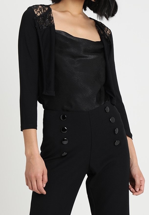 Femme portant un haut en satin noir sans manches, un cardigan court noir avec des épaules en dentelle, et un pantalon noir taille haute avec deux rangées de boutons.