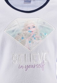 Sudadera de forro polar en gris claro con un parche bordado en forma de diamante que presenta un personaje y lentejuelas. El texto dice: "CREE en ti mismo."