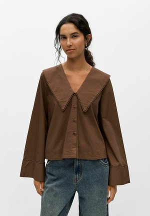 Femme aux cheveux foncés portant un chemisier marron ample à large col et manches évasées, associé à un jean bleu sur un fond blanc uni.