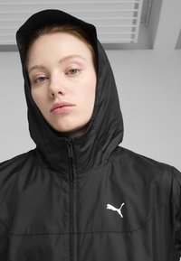Veste zippée noire avec capuche, arborant un logo Puma blanc sur la poitrine gauche. Texture lisse et design minimaliste.