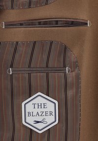 Braunes Sakko mit gestreiftem Taschenfutter, versehen mit einem sechseckigen Aufnäher mit dem Text "THE BLAZER" und einer Scherengrafik. Zwei sichtbare Reißverschlusstaschen.