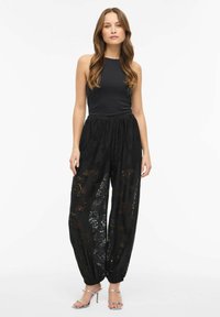 Czarne koronkowe spodnie z kwiatowymi wzorami, elastycznym paskiem w talii i zwężanymi mankietami, zestawione z bezrękawnym czarnym topem i srebrnymi sandałami na obcasie.