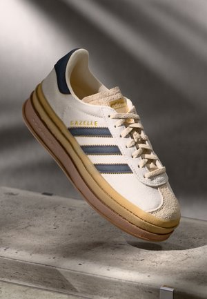 Baskets crème et marine avec trois rayures bleues, accents en daim texturé, semelle épaisse et branding "Gazelle" en doré.