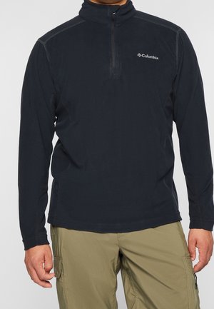 Homme portant un pull polaire noir Columbia à fermeture éclair quart-de-tour et un pantalon cargo kaki, debout devant un fond uni.