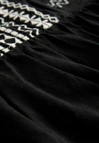 Svart tyg med vita broderade geometriska mönster. Har en rynkad struktur, slät yta och subtil lyster. Idealisk för kläder.