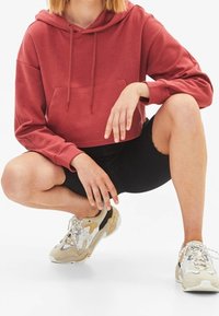 Felpa rossa tagliata con tasca frontale, abbinata a pantaloni corti neri e sneakers beije con dettagli in rete e gomma. Design semplice.
