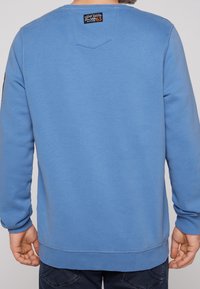 Hellblaues Sweatshirt aus Baumwollmischung mit gerippten Bündchen und Saum. Verfügt über ein Etikett auf der Rückseite und ein dreieckiges Detail unterhalb des Halsausschnitts.
