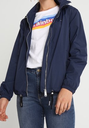 Kvinde iført en marineblå lynlåstrøje over en hvid T-shirt med et regnbuegrafik, kombineret med blå denimjeans.
