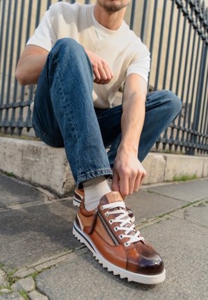 Homme portant une chemise blanche et un gilet, jean bleu, assis sur le trottoir en train de lacer des baskets en cuir marron avec des lacets blancs et un détail zip.
