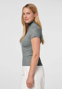 Grijze geribbelde coltrui met korte mouwen, aansluitend ontwerp en gestructureerde stof, gecombineerd met witte broek tegen een neutrale achtergrond.