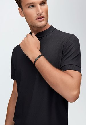 Black short-sleeve polo shirt featuring a textured weave, high collar, and ribbed detailing. A slim black bracelet adorns the wrist.