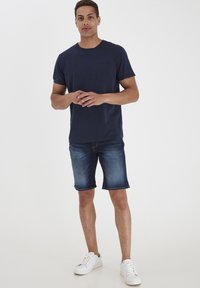 T-shirt iz mornarsko modrega bombaža s žepom na prsih, kombiniran z denim kratkimi hlačami in belimi teniskami. Ohlapen kroj in gladka tekstura.