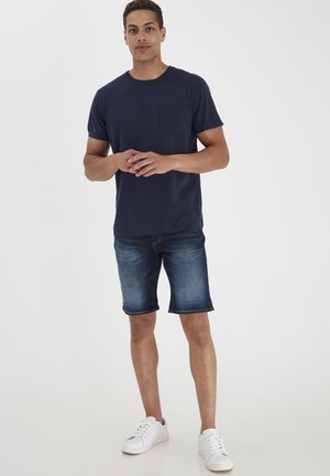 T-shirt iz mornarsko modrega bombaža s žepom na prsih, kombiniran z denim kratkimi hlačami in belimi teniskami. Ohlapen kroj in gladka tekstura.