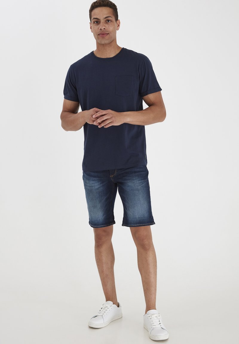 T-shirt iz mornarsko modrega bombaža s žepom na prsih, kombiniran z denim kratkimi hlačami in belimi teniskami. Ohlapen kroj in gladka tekstura.