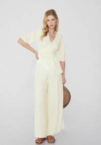 Top envolvente de color amarillo claro, texturizado, con mangas anchas, combinado con pantalones sueltos de rayas y sandalias marrones con flecos. Sostiene un sombrero de paja.