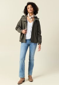 Giacca leggera verde oliva con cappuccio, polsini elastici e tasche frontali. Indossata sopra una maglietta bianca a collo a V e jeans blu, abbinata a stivali beige.