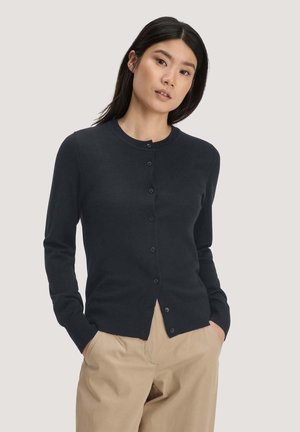 Frau mit langen dunklen Haaren, die eine schwarze, geknöpfte Strickjacke und beige Hosen trägt, steht mit den Händen in den Taschen vor einem schlichten hellen Hintergrund.