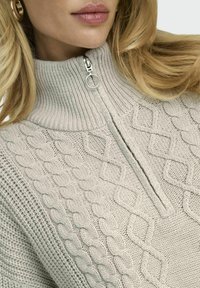 Helles graues Zip-Up-Sweatshirt mit hohem Kragen, das strukturierte Zopfmuster und einen silbernen Reißverschluss mit einem runden Ziehgriff aufweist.