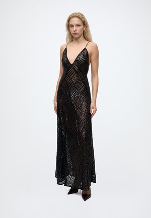 Femme blonde portant une longue robe noire transparente avec des motifs en sequins et des bretelles fines, debout devant un fond clair uni.