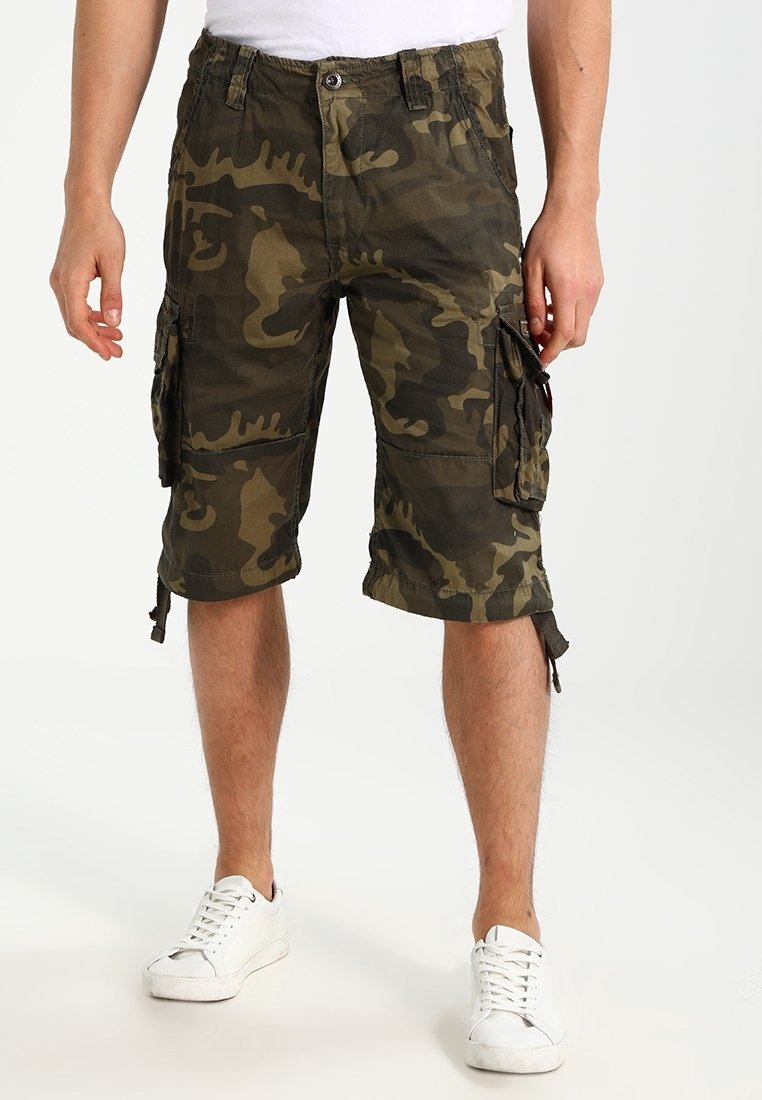 Homme portant un short cargo camouflage vert et marron longueur genou avec plusieurs poches et des baskets blanches.
