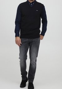 Granatowy sweter z dekoltem w serek, nałożony na niebieską koszulę z kołnierzykiem, w połączeniu z szarymi dżinsami. Na piersi małe logo. Gładka faktura.