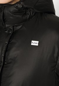 Sort puffet jakke med en glansfuld overflade, der har hætte, trykknapper og et hvidt Levis-logo patch på venstre bryst.