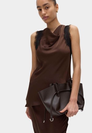 Vrouw draagt een mouwloos bruin satijnen topje met zwarte kanten rand, houdt een donkerbruine leren handtas vast tegen een effen achtergrond.