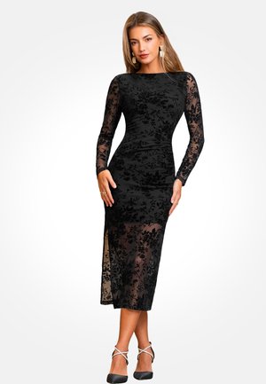 Robe noire à manches longues avec une superposition en dentelle florale transparente, silhouette ajustée, fente latérale et ourlet arrivant au genou. Associée à des talons noirs.