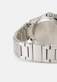 Edelstahl-Uhr mit einem metallischen Armband, das ein rundes Gehäuse und eine polierte Oberfläche aufweist. Auffälliges, geprägtes Logo auf dem Verschluss.
