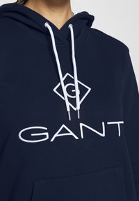 Marineblauwe sweatshirt met capuchon en witte koordjes, groot wit "GANT" logo en een buidelzak aan de voorkant. Zachte textuur.